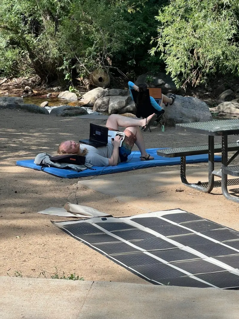 Someone lying on a blue mat by a creek with a laptop on their chest and a keyboard in hand, a solar panel unrolled in the foreground