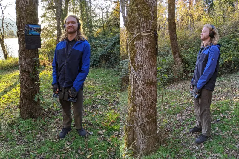 A laptop strapped to a tree trunk with rope, its user standing beside it holding a split keyboard